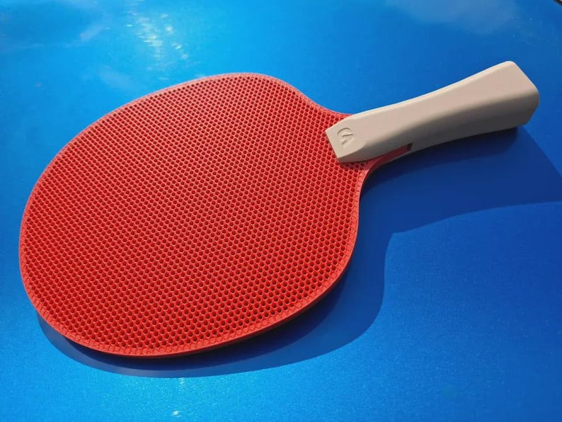 May include: A red ping pong paddle with a textured surface and a light gray handle. The paddle is set against a blue background, with a shadow cast to the side. The handle has a small logo.