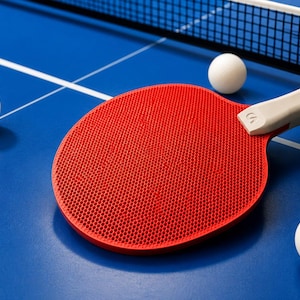 May include: A red ping pong paddle with a white handle rests on a blue table, next to three white ping pong balls. A white net is visible in the background. The table has white lines.