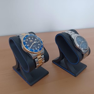 Può includere: Due orologi da polso esposti su supporti neri. Un orologio ha un cinturino oro e argento con una lunetta blu e oro. L'altro orologio ha un cinturino argentato e un quadrante blu. I supporti sono neri con una parte superiore curva.