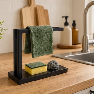 Può includere: Un portasciugamani da cucina in metallo nero con un asciugamano verde, una spugna gialla e una spugna in lana d'acciaio. Il supporto è su un piano di lavoro in legno vicino a un lavandino e una pianta.