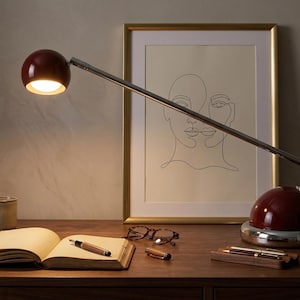 May include: A burgundy desk lamp with a chrome arm and base illuminates an open book and a pen. A framed line drawing of two faces and a mug are also on the wooden desk. Eyeglasses and a pen holder are also visible.