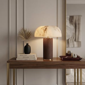May include: A modern table lamp with a marble-like shade and a woven brown base sits on a wooden console table. A black vase with a dried palm frond and stacked books are also on the table. A mirror reflects the room.