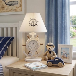 May include: A white table lamp featuring a ship wheel base with an integrated analogue clock and a white lampshade decorated with a black ship wheel graphic. The lamp base is accented with a rope detail and a small anchor at the bottom.