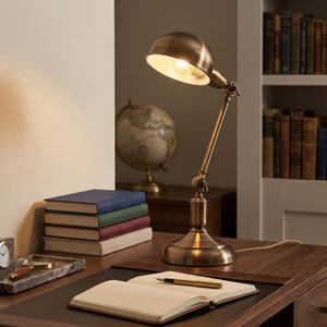 Puede incluir: Una lámpara de escritorio de latón antiguo con un brazo ajustable y una pantalla en forma de cúpula. La lámpara está encendida, proyectando un brillo cálido. Libros, un globo terráqueo y un cuaderno con un bolígrafo también están en el escritorio.