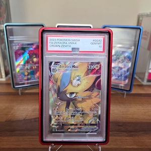 Op de afbeelding: Een gegradeerde 2023 Pokemon SWSH Crown Zenith Zeraora VMAX ruilkaart in een rode plastic houder. Het label vermeldt 2023 POKEMON SWSH FA/ZERAORA VMAX CROWN ZENITH #GG42 GEM MT 10. De kaart toont een geel en zwart personage met 320 HP.