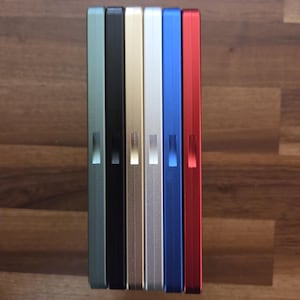 May include: A row of seven rectangular, metallic cases in various colors: green, black, gold, silver, blue, and red. Each case has a small, rectangular indentation on the side. The cases are arranged vertically on a wooden surface.