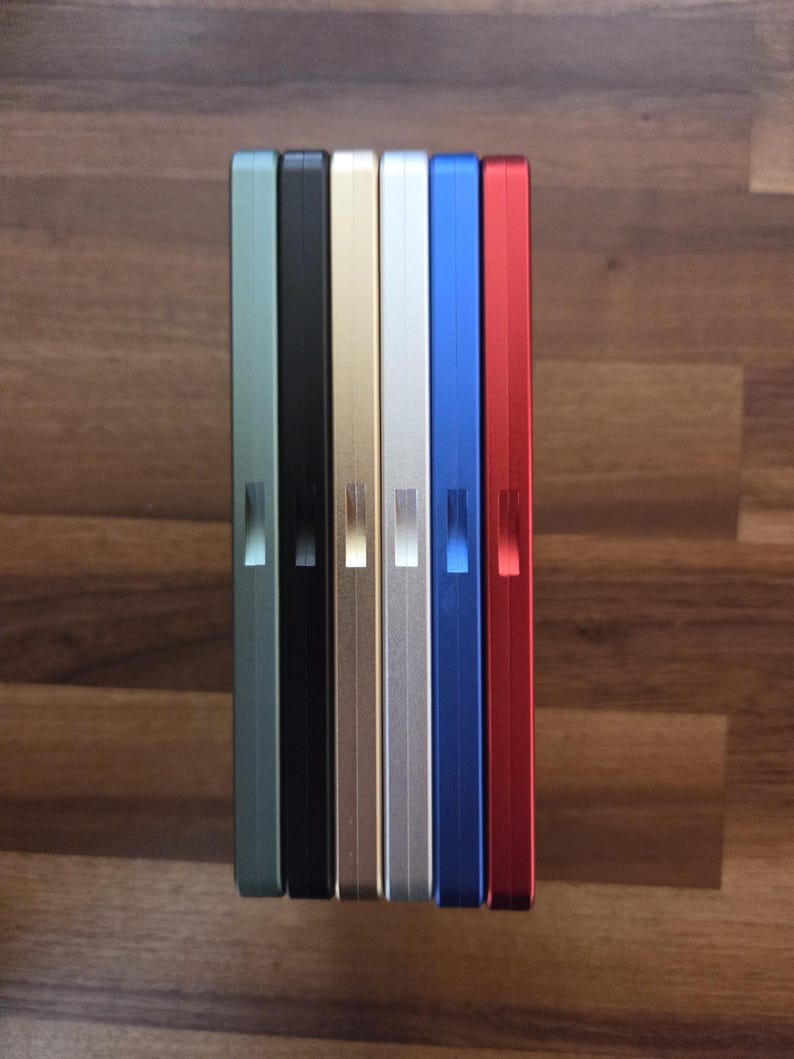 May include: A row of seven rectangular, metallic cases in various colors: green, black, gold, silver, blue, and red. Each case has a small, rectangular indentation on the side. The cases are arranged vertically on a wooden surface.