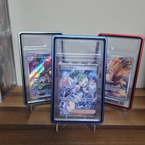 May include: Three trading cards in protective cases. The center card features a character named Lisia's Appeal, with the word "TRAINER" above the character. The cases have blue, red, and silver frames. The cards are displayed on clear stands.