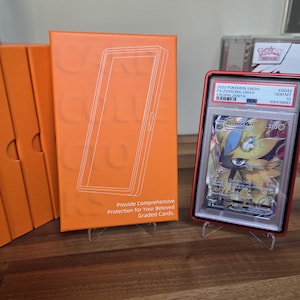 May include: An orange card holder with a white outline of a card. A graded Pok&eacute;mon card in a red case is displayed on a stand. The card features a Zeraora VMAX character. Text on the holder reads, "Provide Comprehensive Protection for Your Beloved Graded Cards."