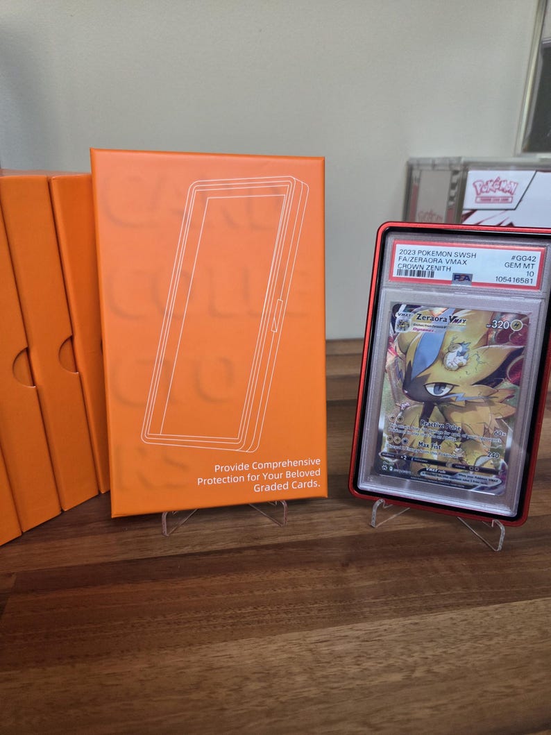 May include: An orange card holder with a white outline of a card. A graded Pok&eacute;mon card in a red case is displayed on a stand. The card features a Zeraora VMAX character. Text on the holder reads, "Provide Comprehensive Protection for Your Beloved Graded Cards."