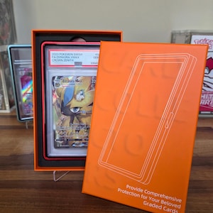 May include: An orange box with a graded Pok&eacute;mon card inside. The card features a Zeraora VMAX character. The box has the text "Provide Comprehensive Protection for Your Beloved Graded Cards."