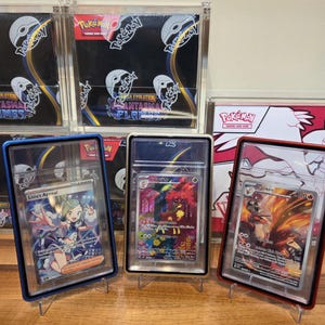 May include: Three Pok&eacute;mon trading cards in protective cases, each displayed on a clear stand. The cards feature colorful illustrations and text, including "Lisia's Appeal" and "Blaziken". Background includes unopened Pok&eacute;mon card packs.