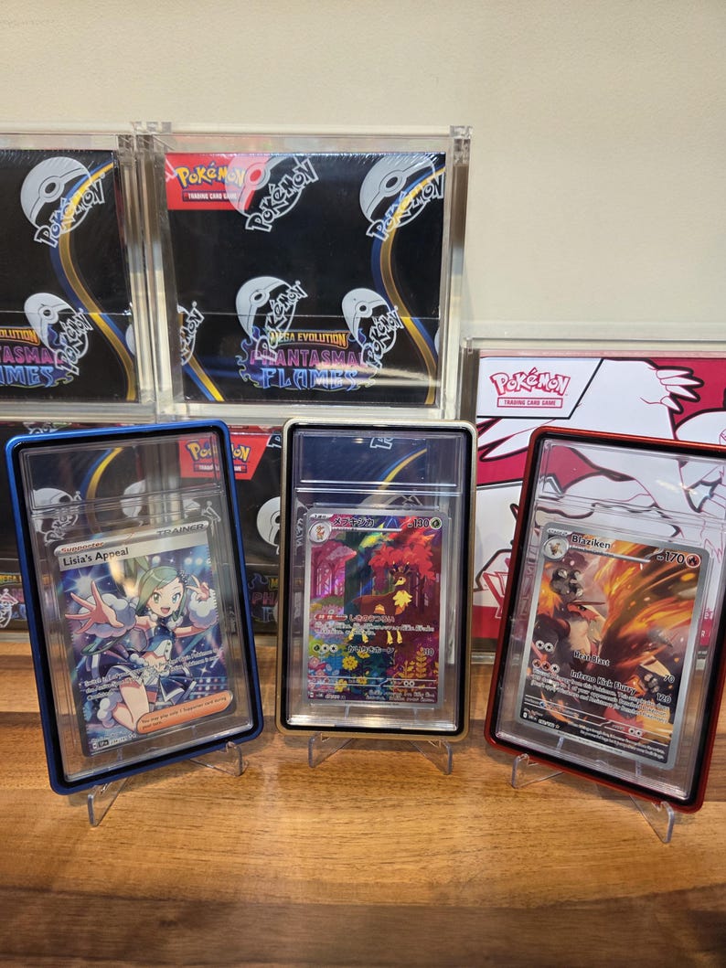 May include: Three Pok&eacute;mon trading cards in protective cases, each displayed on a clear stand. The cards feature colorful illustrations and text, including "Lisia's Appeal" and "Blaziken". Background includes unopened Pok&eacute;mon card packs.