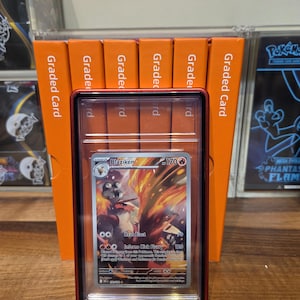 May include: A Blaziken Pok&eacute;mon card in a graded case, featuring a fiery illustration. The card is encased in a red-bordered holder, with orange graded card holders in the background. The card displays the text "Heat Blast" and "Inferno Kick Flurry".
