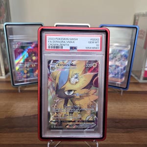 May include: A graded Pok&eacute;mon card featuring Zeraora VMAX from the Crown Zenith set. The card is encased in a red holder with the text "2023 POKEMON SWSH FA/ZERAORA VMAX CROWN ZENITH" and a grade of GEM MT 10. Other cards are visible in the background.