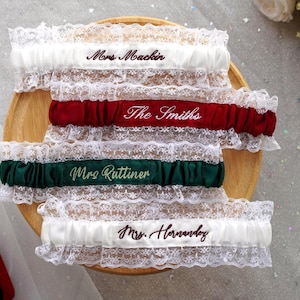 May include: Four bridal garters with lace trim and personalized text. The garters are white, red, and green, with embroidered names in coordinating colors. They are displayed on a wooden plate, with a red ring box in the corner.