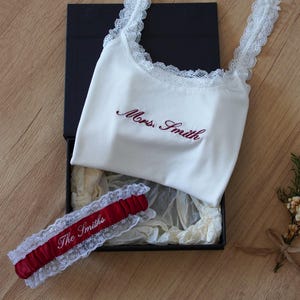 Könnte beinhalten: Ein weißes geripptes Tanktop mit Spitzenbesatz und dem gestickten Text Mrs. Smith in Dunkelrot, kombiniert mit einem passenden dunkelroten Satin-Strumpfband mit weißem Spitzenbesatz und dem gestickten Text The Smiths, in einer schwarzen Box.