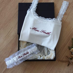 May include: A white ribbed camisole with lace trim and the embroidered text Mrs. Smith, paired with a matching white lace garter featuring the embroidered text Mrs. Shields, both presented inside a black gift box with tissue paper.