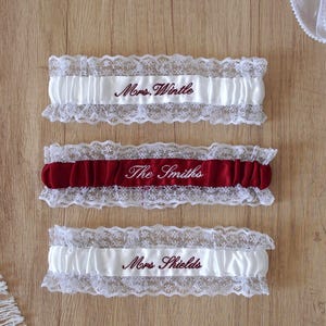May include: Three elasticized garters featuring white lace trim. The top and bottom garters are white satin with dark red embroidery reading Mrs. Wintle and Mrs. Shields. The middle garter is deep red satin with white embroidery reading The Smiths.