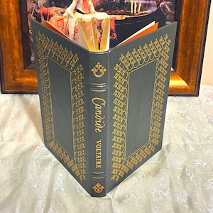 May include: A dark gray book with gold decorative accents and the title "Candide Voltaire" in gold lettering. The book is open, revealing gold-edged pages. The background includes a framed painting.