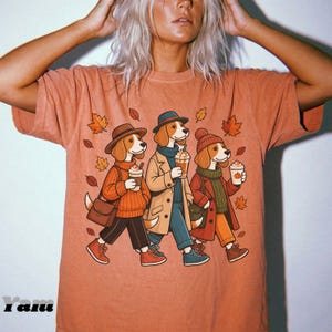 Puede incluir: Una camiseta de color terracota con una ilustración gráfica de tres perros beagle vestidos con ropa de otoño, con sombreros y tazas de café. El diseño incluye hojas naranjas cayendo y el texto "Yam" en la esquina inferior izquierda.