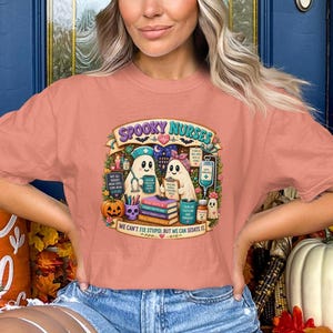 Puede incluir: Una camiseta color melocotón con un gráfico de dos fantasmas vestidos de enfermeros con el texto "SPOOKY NURSES" y "WE CAN'T FIX STUPID, BUT WE CAN SEDATE IT." El diseño incluye suministros médicos, libros, una calabaza y murciélagos.