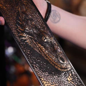 May include: A black leather strap featuring a detailed dragon head design. The dragon's eye is gold, and the scales are intricately carved. The strap is held by a person, with a blurred background.