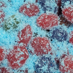 Puede incluir: Primer plano de caramelos coloridos cubiertos de cristales de azúcar azules. Los caramelos son rojos, verdes y azules, creando una apariencia vibrante y texturizada. La imagen destaca el recubrimiento azucarado y la variedad de colores.