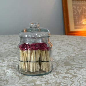 May include: A clear glass jar with a lid, filled with bundles of wooden matches. The match heads are a deep red color. The matches are secured with green rubber bands. The jar sits on a patterned tablecloth.