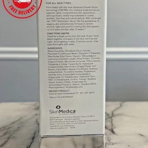 May include: A white rectangular box of SkinMedica TNS Advanced+ Serum. The front features black text detailing product benefits, directions, and ingredients. A silver metallic base is at the bottom. A red circular free delivery stamp is in the corner.
