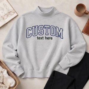 May include: A light gray mock neck sweatshirt featuring the word CUSTOM in large dark blue collegiate-style lettering across the chest, with the words text here embroidered in smaller dark green font directly underneath the main text.
