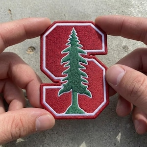 May include: A red embroidered patch in the shape of the letter S with a white border. Centered on the red letter is a green embroidered pine tree graphic. The patch features a dense, textured thread construction with clean, defined edges.