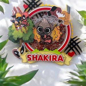 May include: A decorative, layered art piece featuring a cartoon pug with Deadpool and Wolverine characters. The design includes a red and white circular frame, the name "SHAKIRA", and various embellishments. The background is filled with red and green glitter.