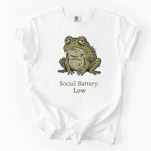 Op de afbeelding: Wit T-shirt met een cartoon illustratie van een chagrijnige groene pad. De tekst "Social Battery: Low" staat onder de pad gedrukt. Het shirt heeft een ronde hals en korte mouwen.
