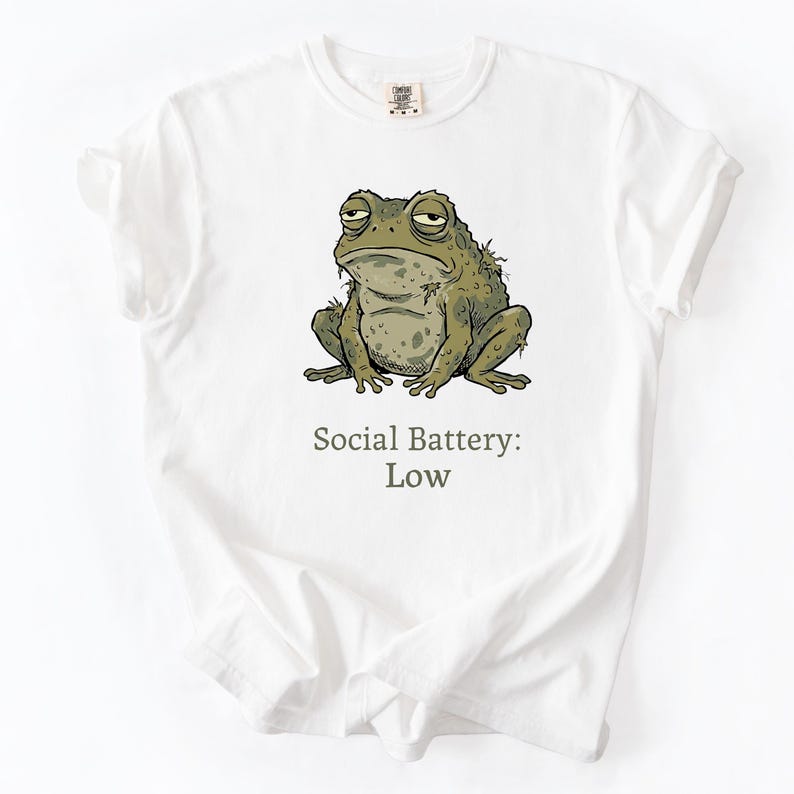 Op de afbeelding: Wit T-shirt met een cartoon illustratie van een chagrijnige groene pad. De tekst "Social Battery: Low" staat onder de pad gedrukt. Het shirt heeft een ronde hals en korte mouwen.