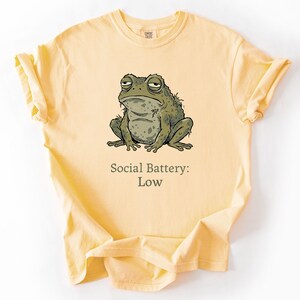 Op de afbeelding: Lichtgeel T-shirt met een cartoon illustratie van een kikker. De kikker is groen en grijs met een vermoeide uitdrukking. Onder de kikker staat de tekst "Social Battery: Low."