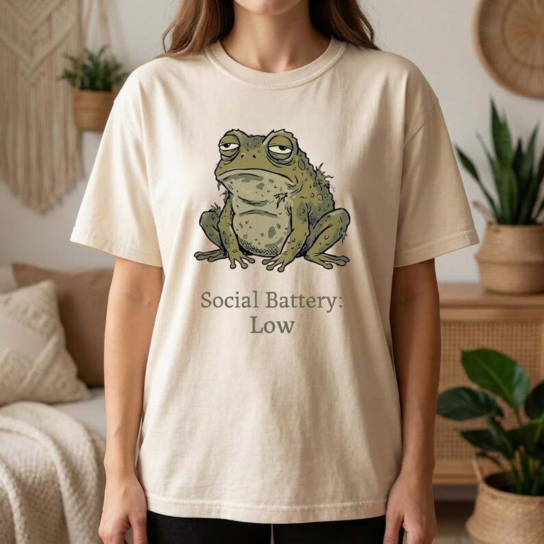 Op de afbeelding: Cr&egrave;mekleurig T-shirt met een illustratie van een chagrijnige groene kikker. De kikker zit met gespreide poten. Onder de kikker staat de tekst "Social Battery: Low".