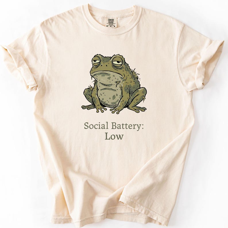 May include: A cream-coloured t-shirt with a cartoon illustration of a grumpy frog. The frog is green and grey, with a tired expression. The text below the frog reads "Social Battery: Low."