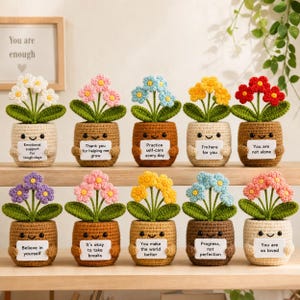 May include: Ten crocheted potted flowers with smiling faces, each holding a white sign with encouraging text. The flowers feature various colors including white, pink, blue, yellow, red, and purple, set in brown or cream-colored yarn pots.