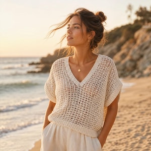 Può includere: Top in maglia color crema con lavorazione traforata, scollo a V e maniche corte. Il top è abbinato a pantaloni colorati chiari. L'immagine è scattata all'aperto su una spiaggia, con il mare e un tramonto sullo sfondo.