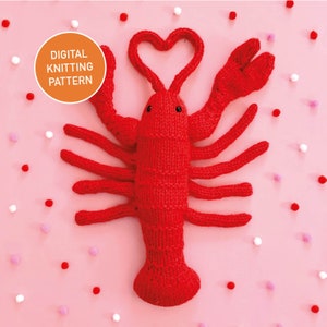 Puede incluir: Un langostino rojo tejido con sus garras levantadas en forma de corazón. El langostino está rodeado de pompones rosas y blancos sobre un fondo rosa. El texto "DIGITAL KNITTING PATTERN" está en un círculo naranja en la parte superior de la imagen.