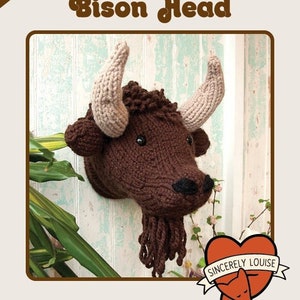 May include: Knit your own Bison Head knitting pattern. The pattern includes instructions for two sizes. The image shows a brown knitted bison head with a beige knitted nose and horns. The bison head is mounted on a wall with a green plant in the background. The image also includes a red heart with the text "Sincerely Louise" and a fox inside the heart.