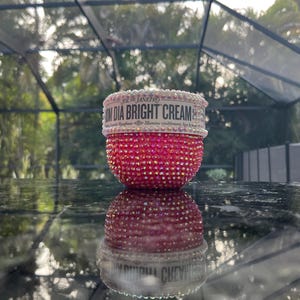 Könnte beinhalten: Ein runder Tiegel der Sol de Janeiro Bom Dia Bright Cream mit einem mit rosa Strasssteinen bedeckten Körper und einem mit weißen Strasssteinen verzierten Deckel. Der Etikettentext lautet Sol de Janeiro Bom Dia Bright Cream.