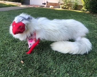 Puede incluir: Un juguete de peluche con forma de zorro, con pelo sintético largo de color blanco, una máscara facial de tela roja y una pata delantera de tela roja. El juguete lleva un pañuelo triangular rosa con un patrón de fresas y pequeñas flores amarillas.