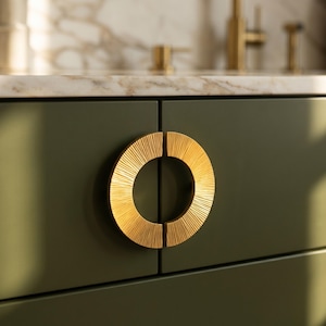 Peut inclure: Gros plan sur une poignée de meuble circulaire dorée avec un design texturé et strié. La poignée est montée sur une porte d'armoire vert foncé. L'arrière-plan présente un comptoir en marbre et des accessoires dorés.