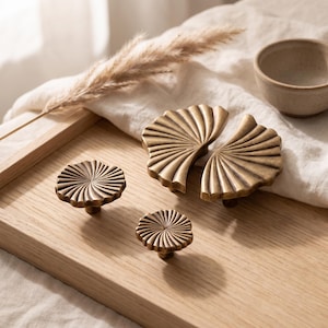 May include: A set of four brass-toned metal cabinet knobs and pulls featuring a radial, fan-like ribbed texture. The collection includes two circular knobs of varying sizes and two semi-circular pulls that form a complete fan shape when joined.