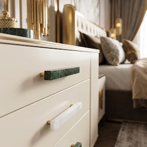 Puede incluir: Una cómoda de color crema con tres cajones horizontales y tiradores rectangulares. Los tiradores superior e inferior son de mármol verde oscuro, mientras que el central es de mármol blanco. Cada uno está fijado con herrajes de metal dorado.