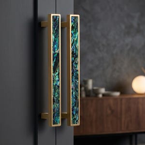 Peut inclure: Deux poignées de porte rectangulaires avec une finition dorée et une incrustation centrale de nacre irisée dans des tons de vert, bleu et noir. Les poignées sont montées sur une porte gris foncé, mettant en valeur un design moderne.