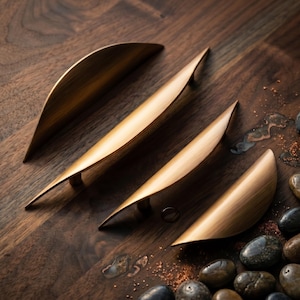 May include: Four curved cabinet pulls with a brushed brass finish. Each handle features a tapered, elongated shape with pointed ends and a smooth, metallic surface. The pulls are arranged in a diagonal line, showcasing their sleek, arching design.