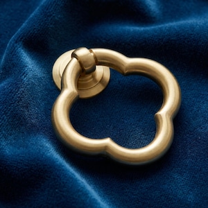 Peut inclure: Une poignée de tirage en métal couleur laiton avec une forme de trèfle à quatre lobes. La quincaillerie présente une finition mate lisse et est fixée à une plaque de montage circulaire. L'anneau articulé pend d'un point de pivot central.